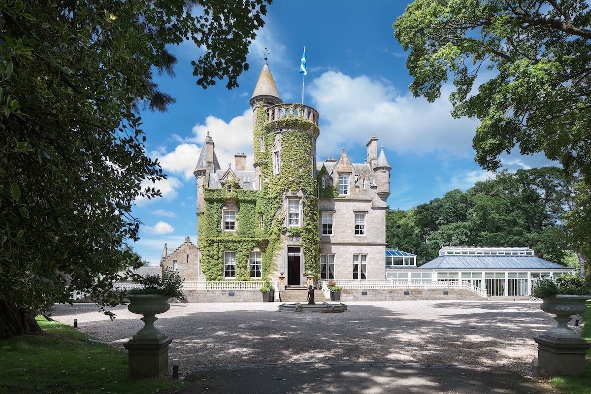 Historic castle with ivy-covered tower, stone facade, and surrounding greenery under a blue sky.